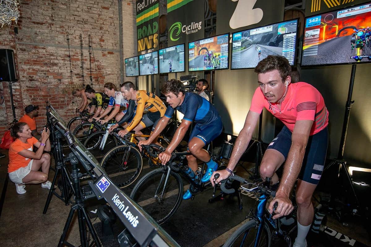 Řada cyklistů šlape na chytrých trenažérech před obrazovkami s virtuální tratí během indoor závodu